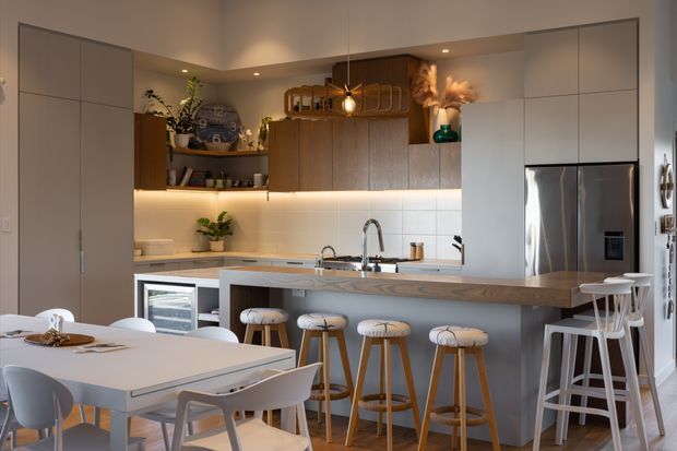 A warmly lit contemporary open-plan kitchen and dining area. The kitchen features pale built-in cabinetry, a long island with a timber-look benchtop, integrated appliances, stainless-steel refrigerator, open timber shelving, pendant lighting, and seating for several people. A dining table with modern chairs occupies the foreground, while decorative plants and accessories add warmth.