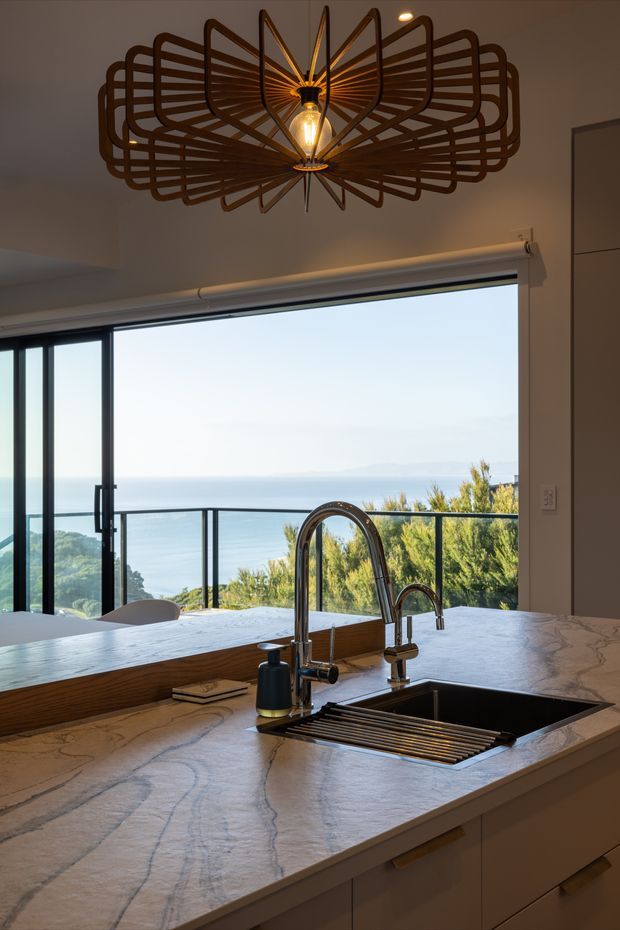 A warmly lit contemporary kitchen focused on a large pale veined stone island with an integrated dark sink, drying rack, soap dispenser, and polished metal gooseneck faucet. Full-height dark-framed glazing and a balcony railing open to a bright ocean and coastal landscape beyond. A distinctive sculptural dark metal pendant hangs above, while light cabinetry runs beneath the island.