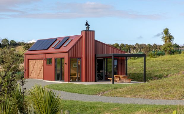 A compact single-storey red corrugated-metal building set in a landscaped rural setting. The structure has a steeply pitched roof with integrated skylights and a chimney, large glazed doors, a covered outdoor terrace with timber bench seating, and solar panels on the roof. Gravel paths and native-style planting surround the building.