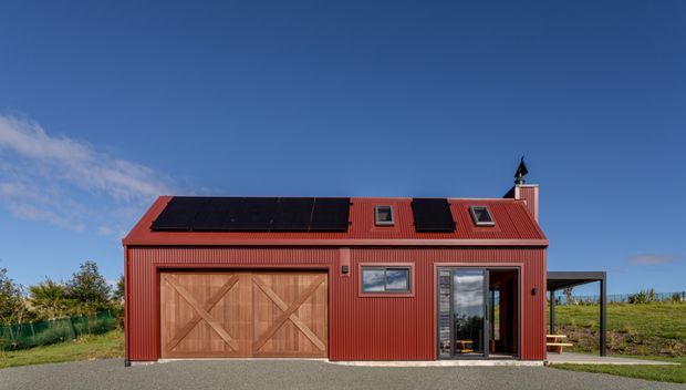 Exterior view of a compact red corrugated-metal building with a steep gabled roof, timber double garage doors, dark solar panels, skylights, chimney, and glazed entry doors. The structure sits in an open rural landscape under a clear blue sky.