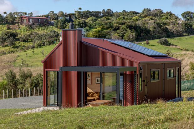 A compact modern rural dwelling set on a gently sloping grassy site, with a dark red corrugated metal facade, asymmetric pitched roof, tall chimney-like vertical volume, and extensive glazed openings. The building appears to contain a small living area visible through the front glazing, with surrounding trees, pasture, and neighbouring hillside vegetation.