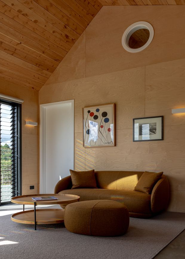 Warm, compact living room with a sculptural brown sofa, matching ottoman, and layered round timber coffee tables. The room has extensive light plywood wall panelling and a sloped timber-lined ceiling, with narrow wall sconces, framed artwork, a high louvered window, and a circular ceiling vent. Natural light creates a cozy, refined atmosphere.