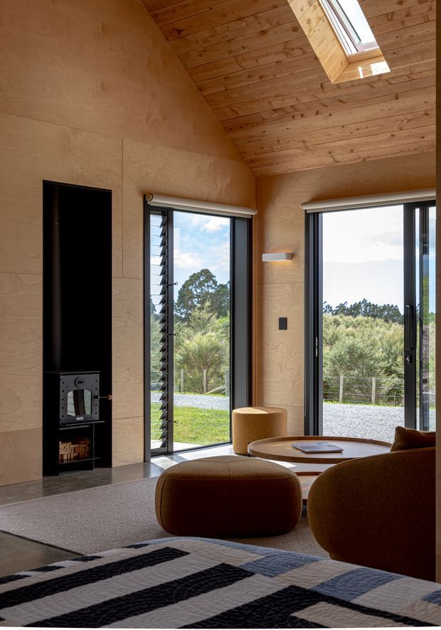 A warm, compact living space with plywood wall and ceiling lining, a steep sloped timber ceiling, skylight, large dark-framed windows and doors, and an integrated black fireplace. Rounded brown lounge seating surrounds a low circular timber table, with a patterned textile in the foreground. The glazing opens to a green rural landscape.