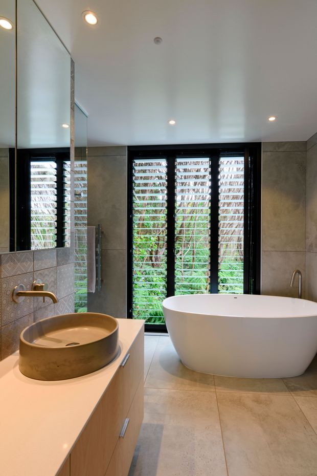 A contemporary bathroom with a freestanding white bathtub positioned beside black-framed louvred windows, a large wall mirror, a round bronze vessel basin, pale built-in vanity cabinetry, and large warm grey stone-look floor and wall tiles. Recessed ceiling lights provide even illumination, while greenery is visible through the windows.