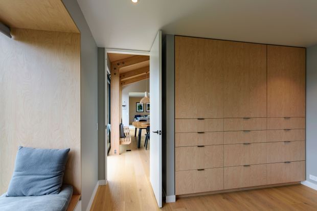 A compact, carefully detailed interior hallway or bedroom storage area with extensive light timber built-in cabinetry and drawers on the right, a timber-lined window seat with a blue cushion on the left, and an open white door leading toward a bright dining or living area. The space features pale timber flooring, grey painted walls, a white ceiling with recessed lighting, and visible timber ceiling beams in the adjoining room. The overall aesthetic is warm, minimal, and contemporary.