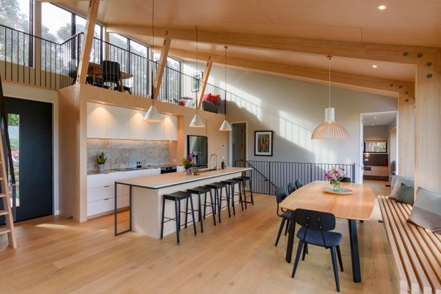 A bright open-plan kitchen and dining area in a contemporary timber-lined home. The space features a large island with seating, white integrated cabinetry, a tiled splashback, pendant lights, timber flooring and ceiling, extensive glazing, a mezzanine level with black balustrades, and built-in timber bench seating around the dining area.