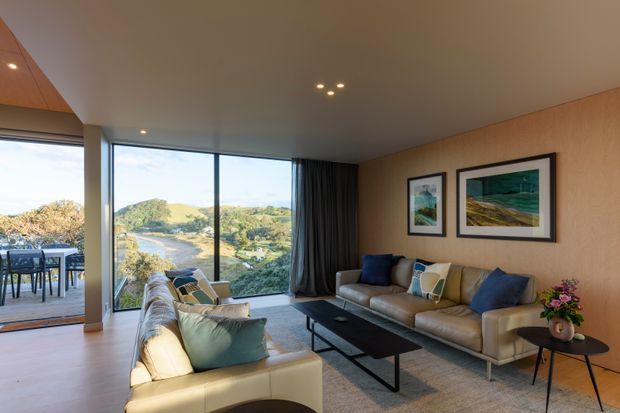 A refined contemporary living room with floor-to-ceiling glazing overlooking a green coastal or hilly landscape. The room features warm timber wall panelling, pale timber flooring, a large neutral sectional sofa and armchair, a black rectangular coffee table, dark curtains, framed artwork, and recessed ceiling lighting. An adjoining outdoor dining terrace is visible through the glazing.