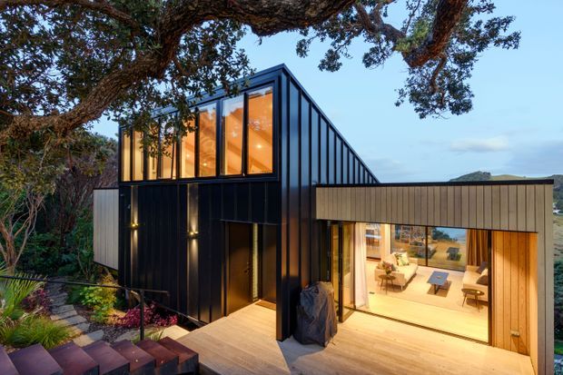 Exterior view of a modern multi-level house set into a landscaped, sloping site. The building features a dark standing-seam metal facade, a strongly pitched roof form with extensive upper-level glazing, and a warm timber-clad ground-floor extension opening onto a broad timber deck. Interior living areas are visible through the large glazed opening, while mature trees and planted garden beds frame the structure.