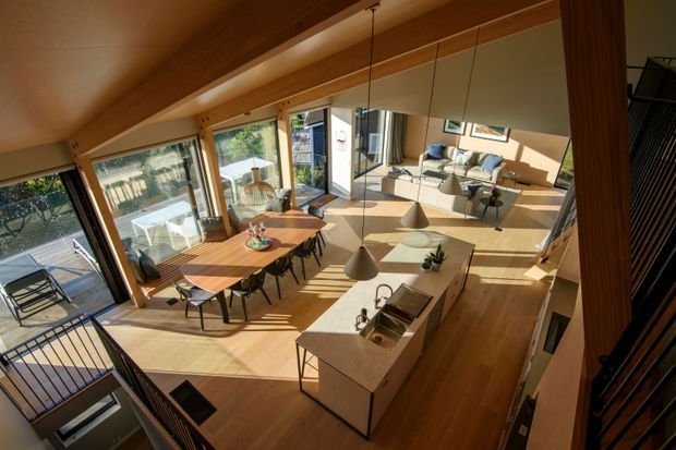 A warm, sunlit open-plan living, dining, and kitchen area viewed from an elevated internal stair or landing. The space features exposed timber beams, extensive glazing and sliding doors, a timber dining table with multiple chairs, a light upholstered sofa, timber flooring, and a central kitchen island with pendant lights. A balcony or exterior deck is visible beyond the glazing, while dark metal stair balustrades frame the foreground.