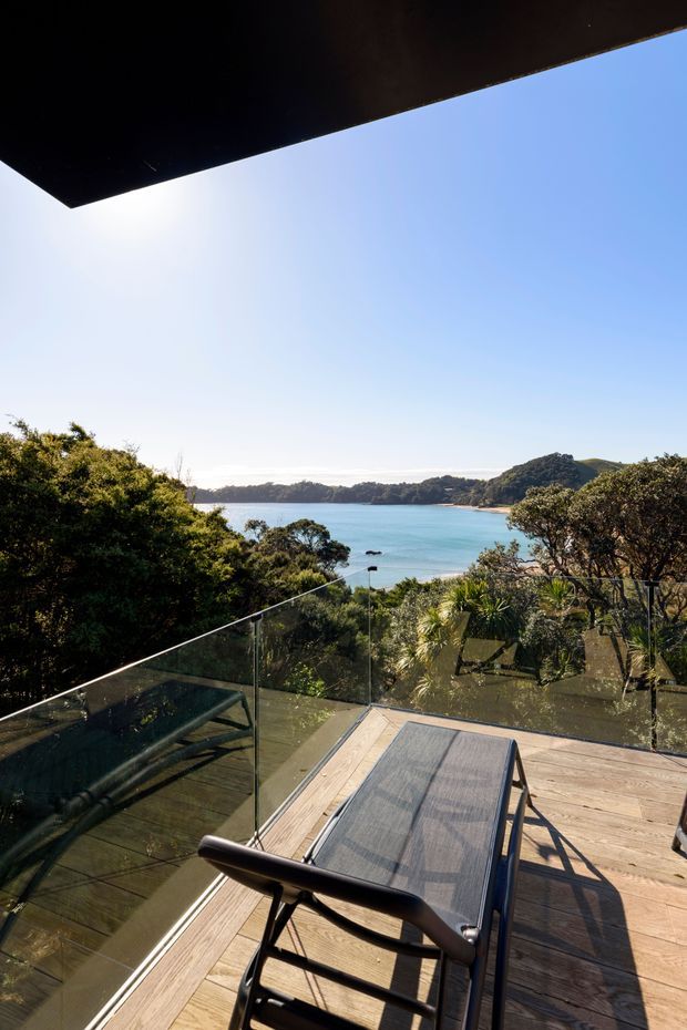 A sunlit elevated timber deck overlooks a turquoise coastal inlet surrounded by dense native vegetation and wooded hills. A clear glass balustrade frames the view, with outdoor lounge chairs in the foreground and a dark projecting roof or overhead eave at the top of the image.