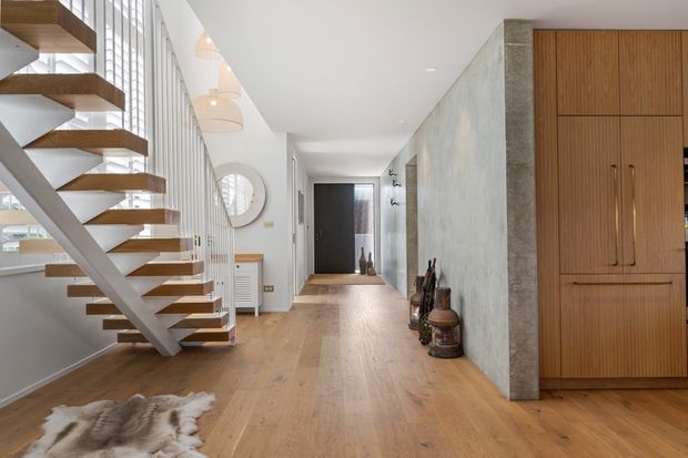 A bright contemporary residential hallway and entry space with a floating timber stair and white structural stringer, expansive vertical glazing, warm timber flooring, a textured grey feature wall, and integrated timber cabinetry. The corridor extends toward a darker interior area, with minimalist pendant lighting and restrained decorative objects.