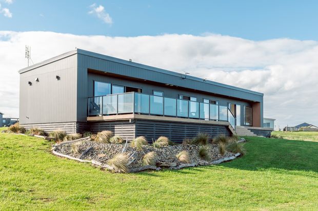 A modern single-storey rectangular house viewed from the front, set in a landscaped grassy site. The building has dark grey-blue corrugated metal cladding, a broad projecting balcony or deck with clear glass balustrades, large glazed doors and windows, and a recessed entry framed in warm timber-toned material. Rock-edged planting beds with ornamental grasses surround the raised deck. The clean linear form and expansive glazing create a contemporary residential exterior.