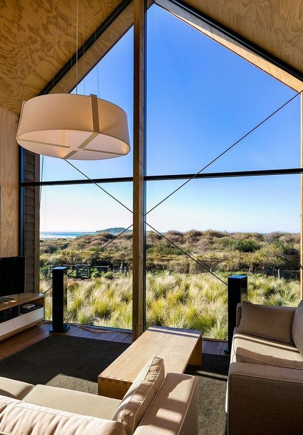 A warm, wood-lined living room with a large sculptural pendant light, extensive floor-to-ceiling glazing, timber-framed windows, and exposed diagonal bracing. Neutral sofas surround a simple timber coffee table, with speakers and media cabinetry along the side. Through the glazing is a coastal dune landscape and ocean horizon.