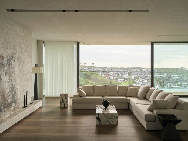 A refined contemporary living room with a large L-shaped beige sectional sofa arranged around a marble cube coffee table. Floor-to-ceiling windows provide expansive urban views, while a textured stone feature wall with a minimalist linear fireplace anchors the room. Dark timber flooring, sheer curtains, recessed linear lighting, and restrained neutral furnishings create a calm, luxurious interior.
