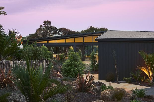 A contemporary single-storey building viewed from a landscaped garden at dusk. The dark vertical-clad wing has a corrugated metal pitched roof, while the glazed central section features warm interior lighting and a long clerestory-like band beneath a shallow sloping roof. Drought-tolerant and native-style planting, rocks, timber edging, and paved paths surround the structure.
