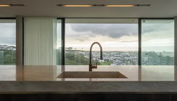 Interior view across a kitchen island toward a wide floor-to-ceiling glazed outlook. A large stone benchtop with an integrated rectangular sink and sculptural gooseneck tap dominates the foreground, with dark fixed cabinetry below. Minimal recessed and linear ceiling lighting, slim dark window frames, and a distant urban/coastal landscape visible beyond the glazing.