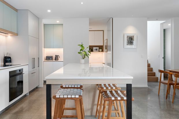 Bright contemporary open-plan kitchen and dining area with a large white island bench, integrated white cabinetry, pale timber accents, black-framed island supports, and woven timber stools. A compact dining area and stairway are visible beyond the kitchen. The space has polished concrete flooring, recessed ceiling lighting, and a clean neutral palette.