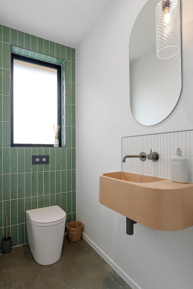 A compact contemporary bathroom featuring a wall-mounted peach-toned basin with exposed brushed-metal tapware, a rounded vertical mirror, a ribbed white splashback, and a green vertically tiled wall surrounding a black-framed window and toilet. The room has a grey floor, white walls, and a sculptural pendant light.