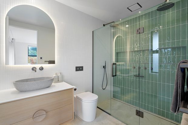 Contemporary bathroom featuring a wall-mounted timber vanity with a round vessel basin, illuminated arched mirror, wall-mounted fixtures, a white toilet, and a large frameless glass shower enclosure. The shower has glossy sage-green vertical tiles, a rainfall showerhead, and handheld shower. White vertically ribbed wall tiles and pale stone-look flooring create a fresh, refined finish.