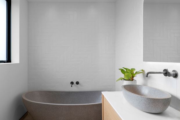 Bright contemporary bathroom featuring a freestanding speckled stone-look bathtub, a matching vessel basin on a light timber vanity, wall-mounted chrome tapware, and white textured geometric wall tiles. A small plant adds a natural accent, while a mirror and window provide additional light and reflection.