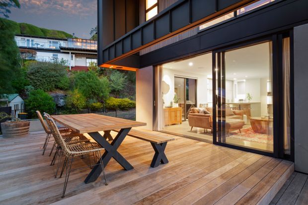 Contemporary residential outdoor entertaining area with a generous timber deck, large timber dining table, woven dining chairs, and a built-in bench beside expansive black-framed sliding glass doors. The doors open to a bright, refined living and dining interior, while landscaped planting and neighbouring residential architecture are visible beyond the deck. The home features strong dark metal framing, warm timber decking, and broad indoor-outdoor connection.