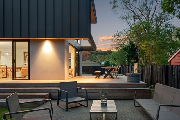 A contemporary home exterior at dusk, viewed from a paved outdoor seating area. The house features dark vertical cladding, smooth light-coloured walls, large glazed openings and a raised timber deck with outdoor dining furniture. A timber privacy fence, garden planting and neighbouring residential structures form the background.