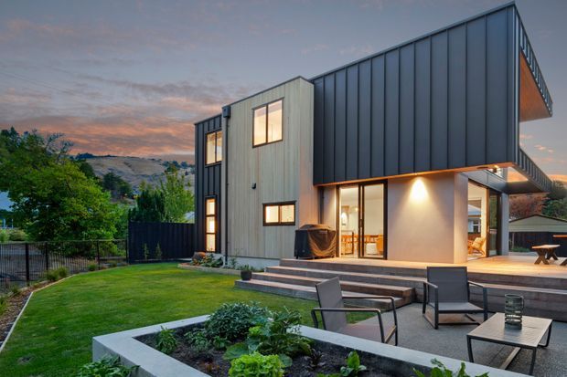 Contemporary multi-level residence with a strong geometric form, dark standing-seam metal cladding, and lighter vertical cladding panels. The home opens onto a raised paved outdoor terrace with lounge furniture and integrated garden beds, overlooking a landscaped yard and distant hills at dusk.