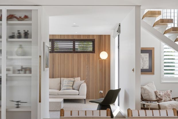 Bright contemporary interior view centred on a compact living room, with a white sofa against a vertical timber feature wall, dark horizontal louvred windows, a spherical pendant light, and a black lounge chair. A glazed display cabinet is visible at left, while an open-riser timber staircase, framed artwork, plantation shutters and additional seating appear to the right.