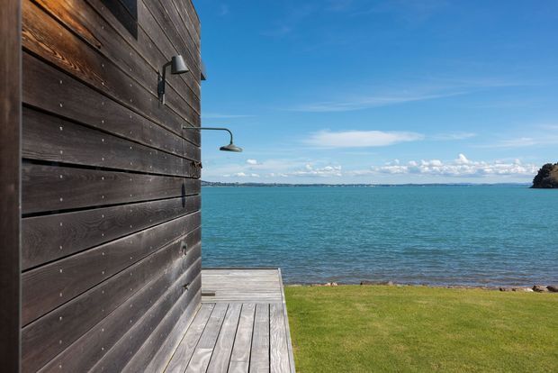 Exterior view of a dark timber-clad building beside turquoise water, with a weathered timber deck, wall-mounted outdoor shower or light fixture, lawn, and expansive coastal horizon under a clear blue sky.