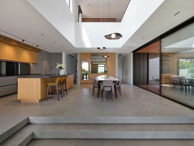 A spacious contemporary open-plan interior with a kitchen on the left and a dining area centred in the room. The space features a large concrete island and flooring, warm timber cabinetry and screening, dark-framed glazed partitions, a broad double-height ceiling void with an upper-level opening, and sculptural pendant lighting. Steps in the foreground create a subtle split-level transition, while a glazed room is visible to the right.