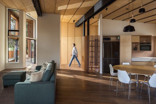 Open-plan contemporary living and dining area with an integrated kitchen. The room features warm timber flooring, a timber-panelled ceiling and walls, full-height glazing, black-framed internal glazing and beams, a dark green sectional sofa, a timber dining table with white chairs, and pendant lighting. A slatted timber screen partially separates the living and kitchen zones.