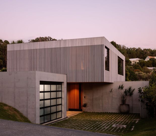 A contemporary two-storey residential house viewed from the street at dusk. The building combines a large smooth concrete lower level with an upper volume screened in closely spaced vertical light-grey battens. A timber front door sits beside a translucent-panel garage door, and a grass-grid driveway leads to the entry. The home has a simple flat-roofed cubic form with large recessed openings and landscaped surroundings.
