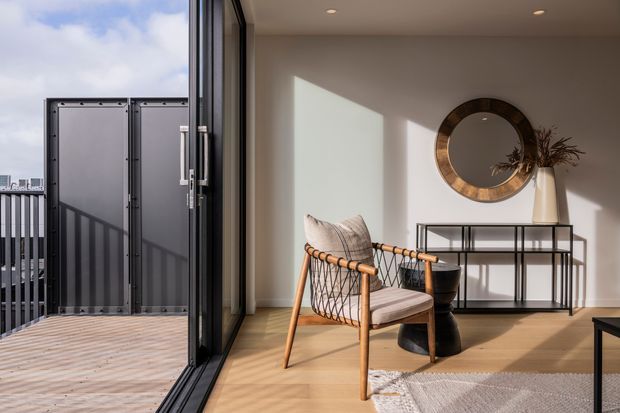 A bright contemporary sitting area opening onto a timber-decked balcony or terrace through full-height black-framed glazing. The room features pale timber flooring, warm white walls, a woven lounge chair, a slim black console table, a round framed wall mirror, a sculptural vase with dried foliage, and recessed ceiling lights. Strong afternoon sunlight creates a calm, warm atmosphere with views toward an urban setting outside.