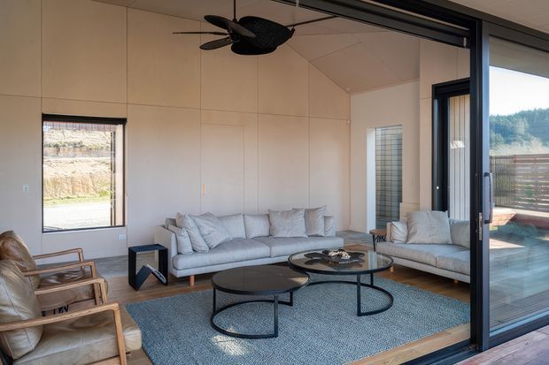 Bright contemporary living room with a neutral palette, pale paneled walls, a sloped ceiling, large sliding glass doors, and views toward an outdoor deck and wooded landscape. The room contains a light grey sofa and armchair, timber-framed lounge chairs, nested round black coffee tables, a textured blue-grey rug, and a dark ceiling fan.