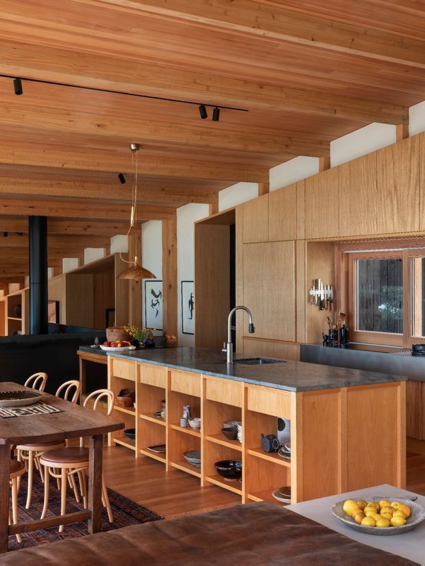Warm, timber-lined open-plan kitchen and dining area with extensive built-in wood cabinetry, a stone island benchtop, integrated sink and tapware, pendant lighting, and a large dining table. The ceiling and walls feature exposed timber lining, with dark accents and artwork adding contrast.