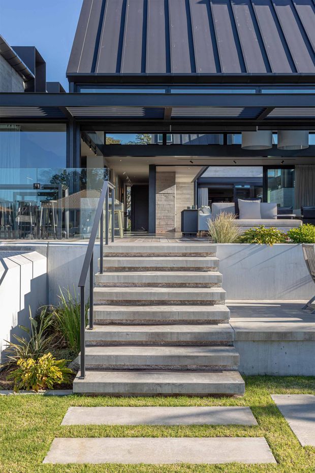 Exterior view of a contemporary multi-level home with a dark metal standing-seam roof and fascia, extensive glazing, a covered outdoor living area, concrete stairs and retaining walls, and landscaped garden edges. The stepped entry leads to a partially furnished outdoor terrace or living space with modern sofas and pendant lights visible beneath the canopy.