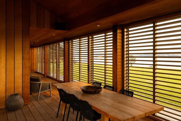 Warm timber-lined dining room or sunroom with a long solid-wood dining table and several dark chairs. Floor-to-ceiling glazing runs along two sides, fitted with horizontal timber louvres or blinds that filter bright outdoor light and views of a grassy landscape. The ceiling, walls, floor, window frames and built-in elements are predominantly timber, creating a strongly tactile, rustic-modern atmosphere.