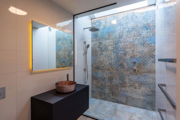Contemporary bathroom featuring a frameless glass shower enclosure, large blue-grey textured feature tiles on the shower walls and floor, pale wall tiles, a wall-mounted dark vanity with a round vessel basin, and a backlit rectangular mirror.