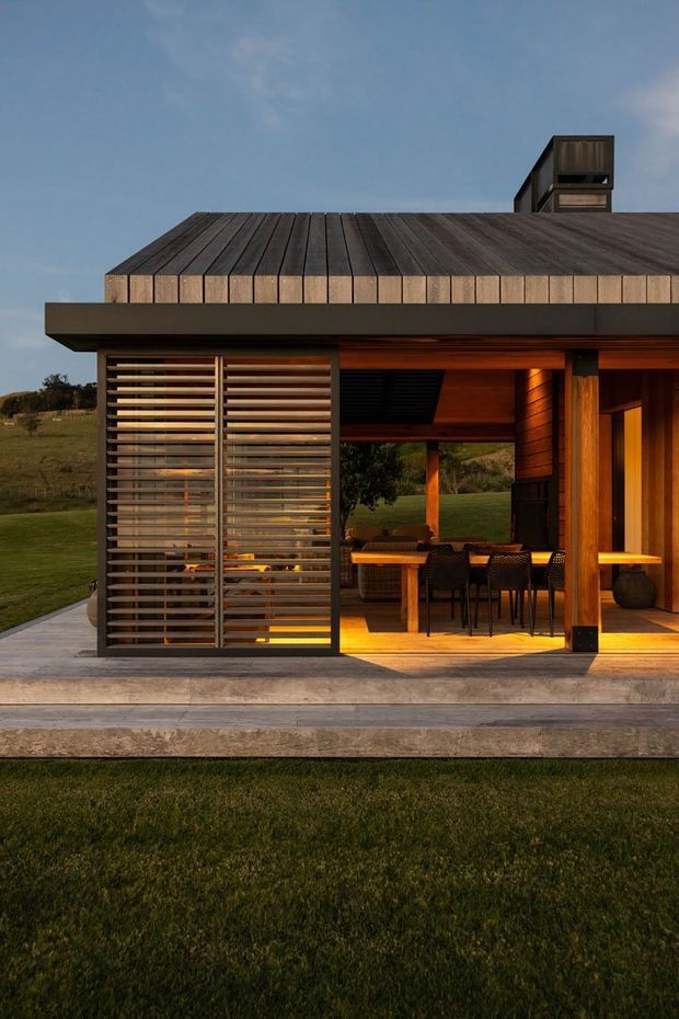 Exterior view of a modern single-storey pavilion or house with a broad low-pitched roof, dark timber roof cladding, timber-framed openings, and horizontal timber screening. A covered outdoor dining area with a table and chairs is visible beneath the deep overhang, opening onto a raised concrete terrace and lawn. Warm interior lighting contrasts with the cool dusk sky and rural landscape.