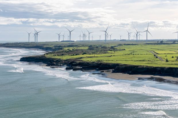 A broad coastal landscape viewed from above, showing waves breaking along a rugged dark-rock shoreline, sandy coves, bright green agricultural fields, and numerous wind turbines across the distant horizon under a cloudy sky.