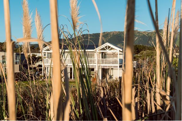 Exterior view of a light-coloured, multi-level residential building with repeated gabled roof forms, white timber-style balcony balustrades, and large glazed openings. The building is partially obscured by tall ornamental grasses and reeds in the foreground, with a wooded hillside visible behind it. Warm late-day sunlight creates a natural, coastal or semi-rural atmosphere.