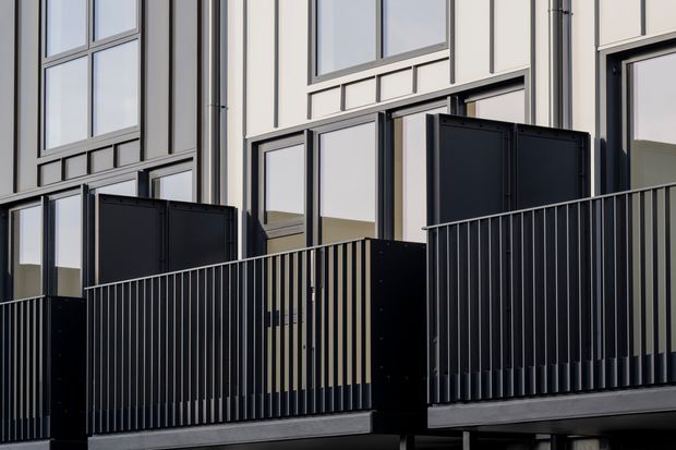 Close-up exterior architectural view of a contemporary multi-level facade with tall glazed openings, pale vertical cladding panels, dark window and door frames, projecting dark balcony platforms, and slim black metal balustrades. The image focuses on the building envelope and balconies, with no clearly identifiable interior room visible.