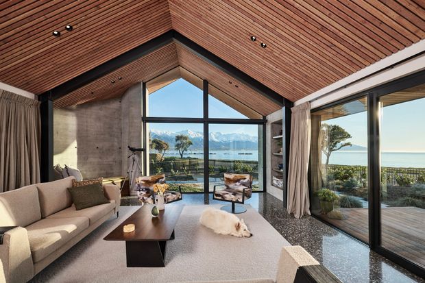 A refined living room with a dramatic pitched timber-lined ceiling, exposed concrete walls, dark-framed floor-to-ceiling glazing, and expansive views toward a lake and mountains. The room features a neutral sofa, dark coffee table, armchairs, a large pale rug, and soft curtains, with an adjoining glazed terrace visible at right.
