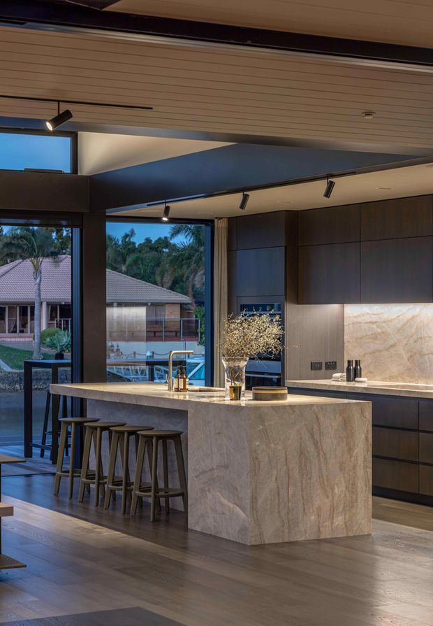 A sophisticated contemporary kitchen with a large stone-clad island and integrated seating for four. Dark timber cabinetry lines the back wall, complemented by a dramatic light-toned stone splashback and benchtop. Floor-to-ceiling glazing opens toward an outdoor setting, while recessed and track lighting illuminate the space. Timber flooring, dark framing, and warm ambient lighting create a refined, luxurious atmosphere.