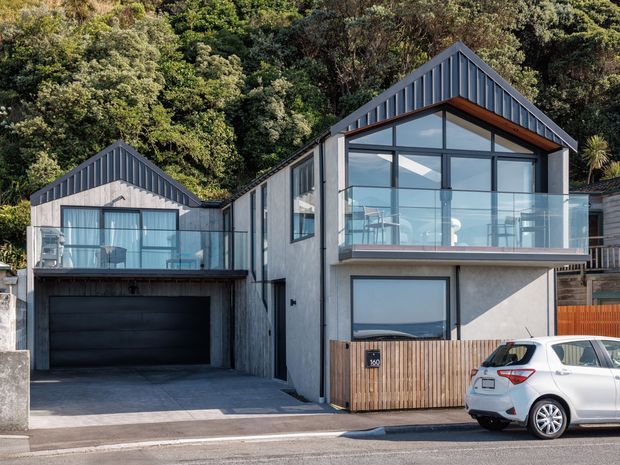 Street-level exterior view of a contemporary multi-level home on a wooded, sloping site. The building has two prominent gabled roof forms, dark vertical metal cladding, light rendered walls, extensive glazing, a glass-balustraded upper balcony, timber screening at the lower level, and an integrated double garage.