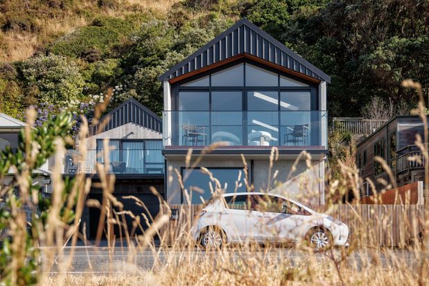 Exterior view of a compact multi-level home set against a wooded, steeply sloping site. The central building has a prominent pitched gable roof, dark vertical cladding, extensive upper-level glazing with visible interior living space, and a balcony or glazed terrace. Adjacent connected volumes and a parked car are visible, with dry grasses in the foreground.
