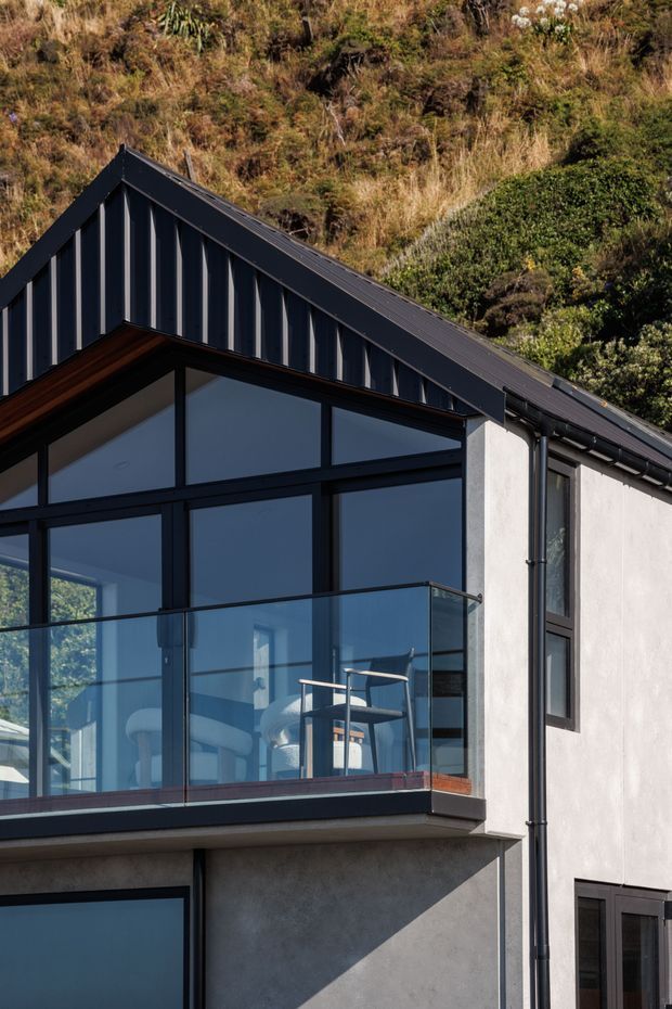 Close-up exterior architectural photograph of a contemporary multi-level dwelling set against a steep vegetated hillside. The building has a dark metal gable roof and matching vertical standing-seam cladding, pale rendered walls, extensive dark-framed glazing, and a projecting balcony with clear glass balustrades and outdoor seating visible behind it.