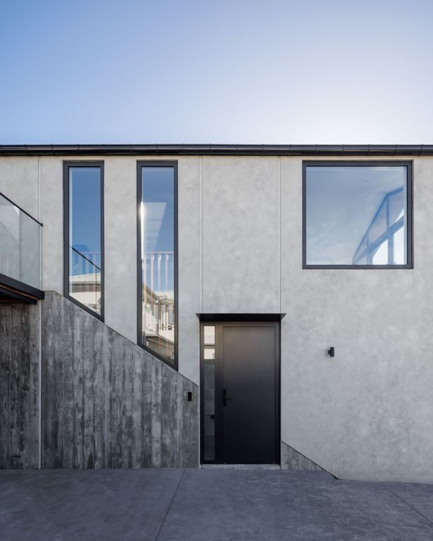 A contemporary residential exterior viewed from a paved approach. The facade combines smooth light-grey rendered panels with dark-framed vertical and square windows, a black entry door with a projecting canopy, and a textured raw concrete retaining wall and stair enclosure. Reflections reveal interior glazing and railings. The composition is minimalist and geometric under a clear sky.
