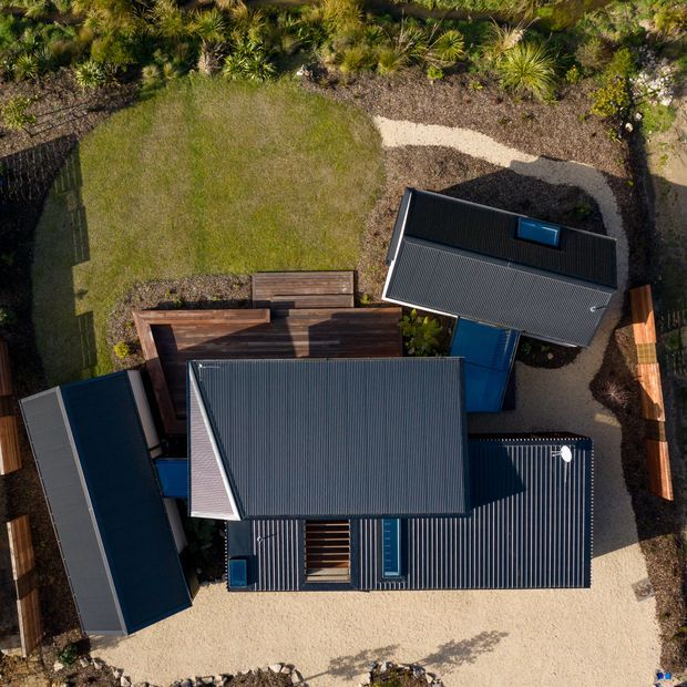Top-down aerial view of a contemporary residential property composed of several dark corrugated metal-roofed structures, connected by timber decking and blue-toned elements. The buildings are arranged around gravel paths, landscaped garden beds, and a lawn, with native-style planting surrounding the site.