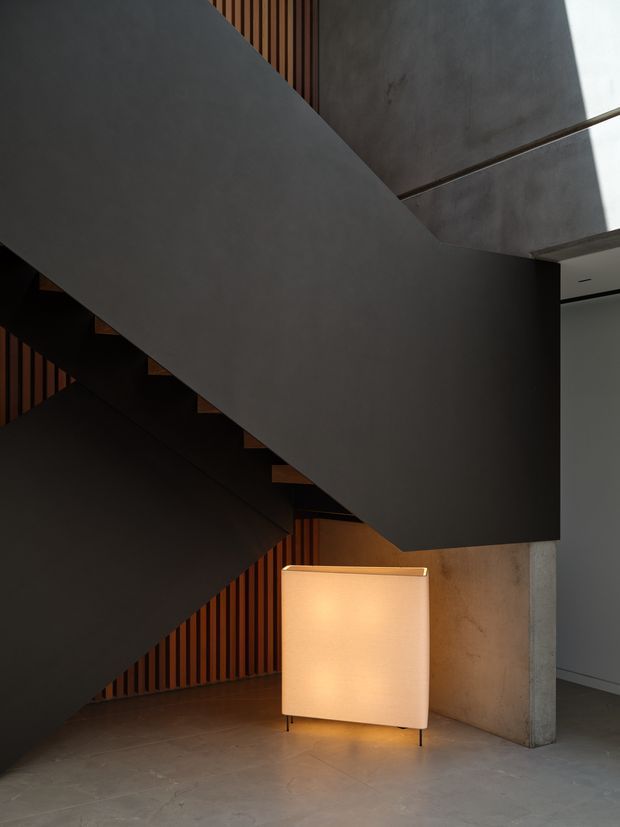 A dramatic, dimly lit interior stairway with a sculptural dark-painted staircase crossing the frame. Vertical timber battens line the stair enclosure, while exposed concrete walls and floor create a monolithic contemporary setting. A softly glowing rectangular floor lamp adds warmth near the base of the stairs, and daylight enters from the upper right.